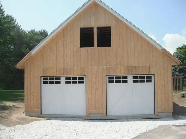 New timber-frame two-car garage with two white windowed doors and a gravel apron