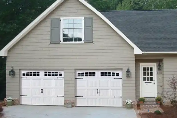 Two white carriage doors on a tan house with shutters, a stoop and planters