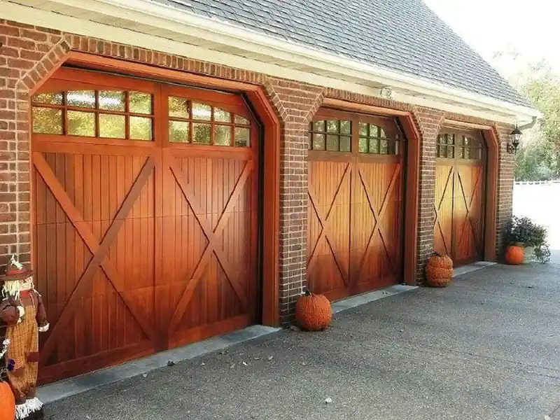 Three stained mahogany X-brace garage doors with window rows on a brick garage, pumpkins by the drive