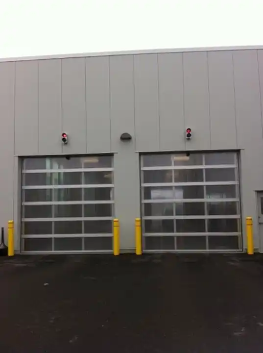 Two full-view glass sectional doors on a grey fire-station-style building with yellow bollards